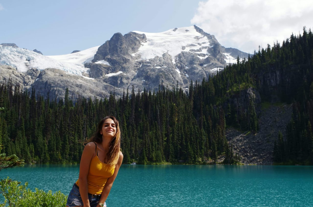 A woman stands near a lake with a mountain in the background.