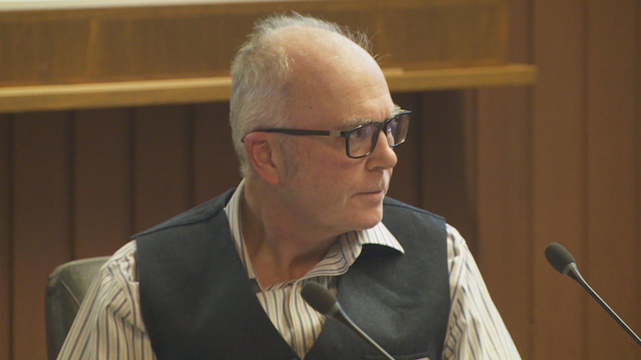 A white man wearing a black waistcoat looks to his left in council chambers.
