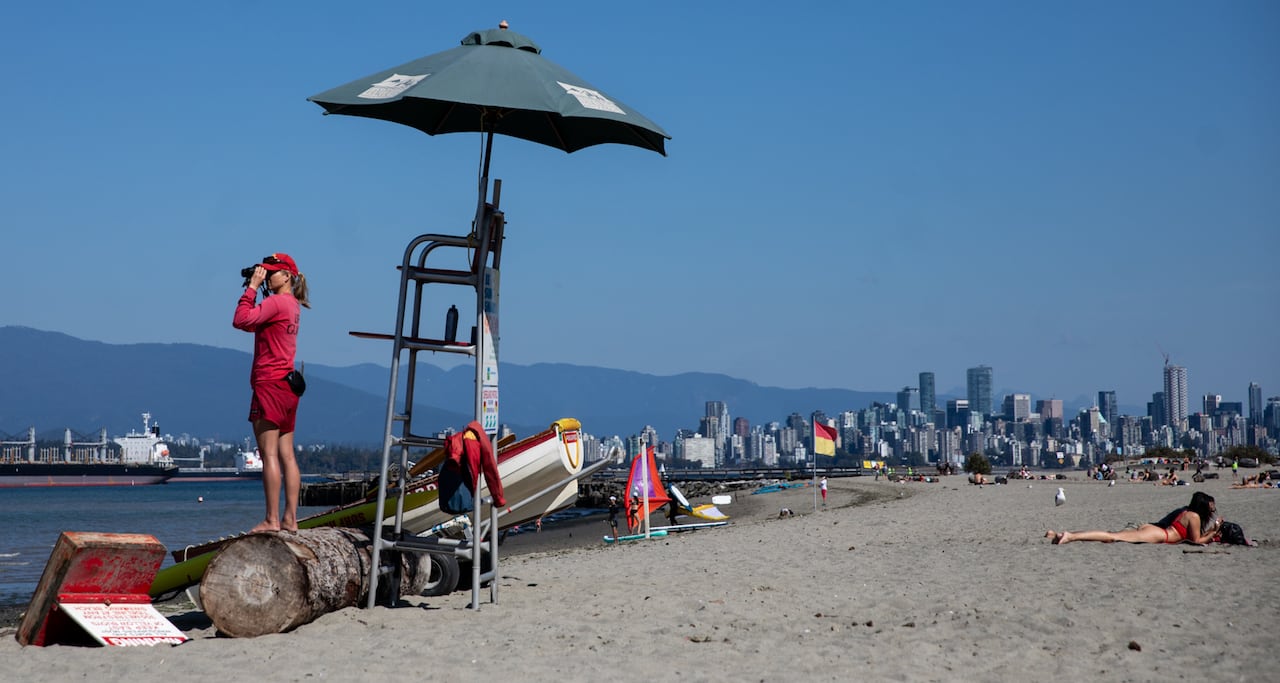A lifeguard looks out using binoculars at a beach while standing on a log, with a downtown skyline visible behind her.