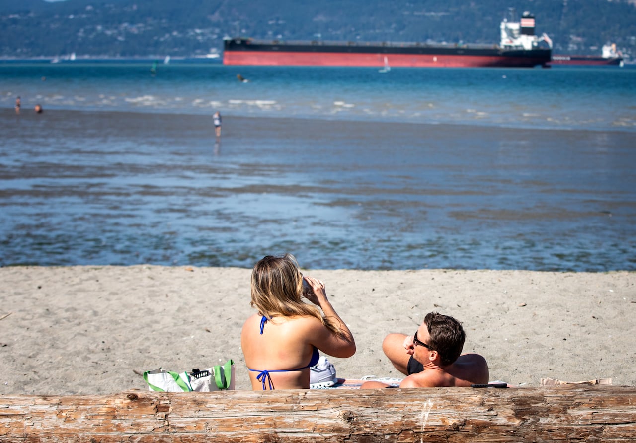 A woman drinks at a beach with a man next to her, and a ship is visible off in the horizon.