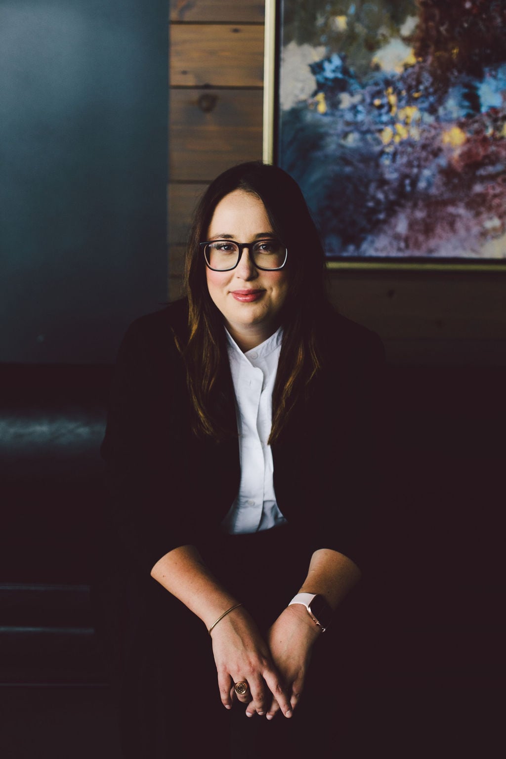 Natasha sits behind a wooden background on a sofa, looking at the camera. 