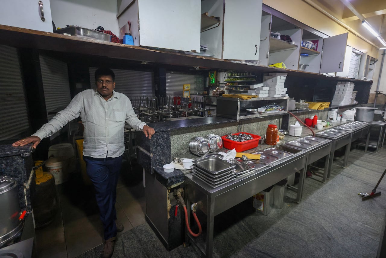 A man in a white shirt and blue pants poses with a serious look on his face as he stands in the kitchen of a restaurant, next to a long stainless steel counter with stainless steel meal trays laid out.
