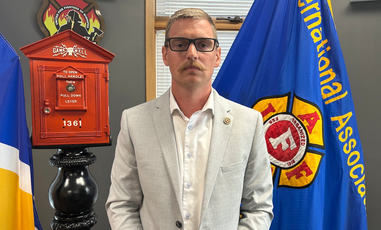 A man wearing a white dress shirt and a grey blazer is pictured standing in front of firefighting-related flags.