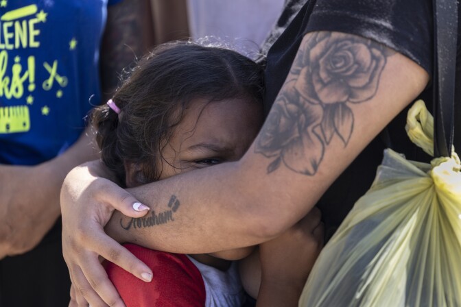 Closeup of a young girl being embraced by a woman who has tattoos on her arm and is wearing a black tshirt.