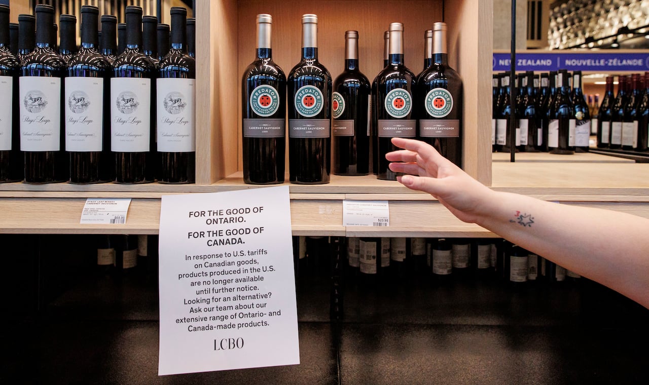 A person's hand reaches toward some wine bottles on a shelf, above a paper that says in part 'products produced in the U.S. are no longer available.'