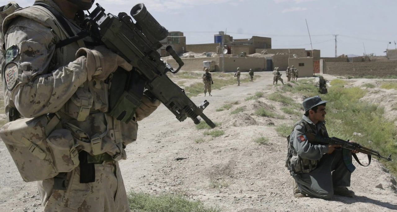 A soldier in desert camoflage holds an assault rifle. There are low, beige buildings in the background