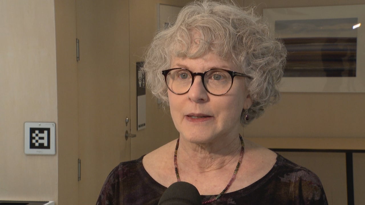 A woman with grey hair and glasses stands in front of a microphone.