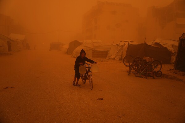 A boy pushes a bicycle carrying jugs of water through a sandstorm in Khan Younis, southern Gaza Strip, Saturday, March 14, 2026. (AP Photo/Abdel Kareem Hana)