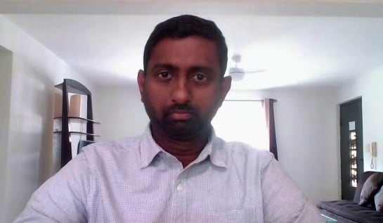 A man wearing a dress shirt sits on a Zoom call in a living room setting.