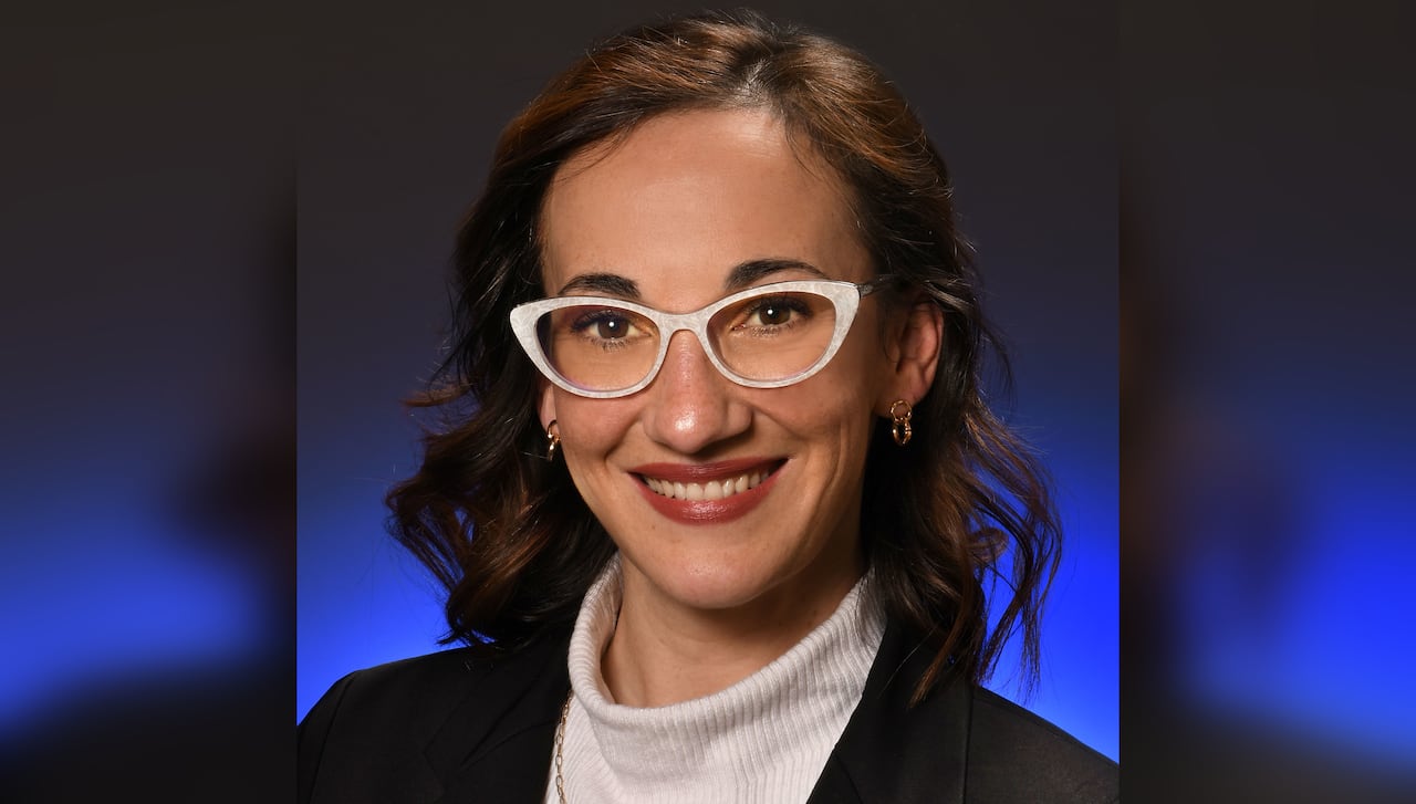 A young woman with shoulder-length brown, curly hair, wearing white cat-eye frame glasses, a white top and black blazer, smiles at the camera.