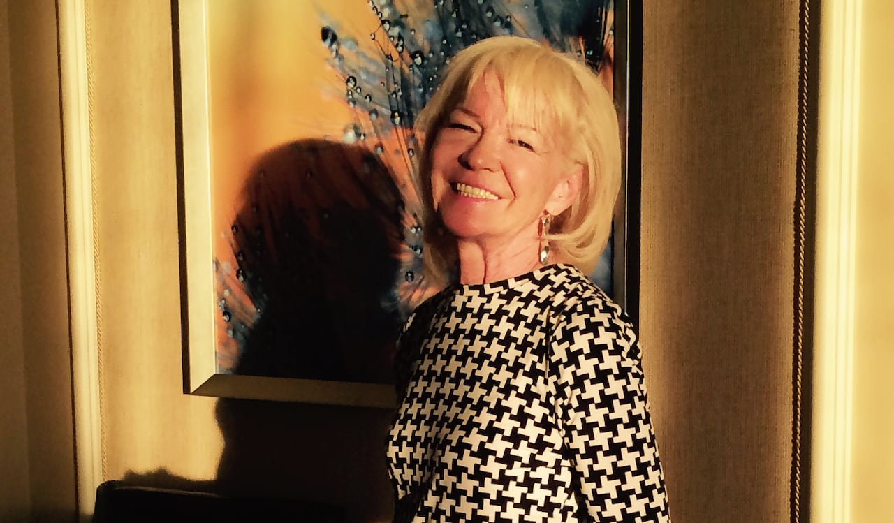 A older white woman, with shoulder-length blonde hair, is wearing a black-and-white dress. She is standing in a condo, evening sunlight shining on her, while she smiles at the camera.