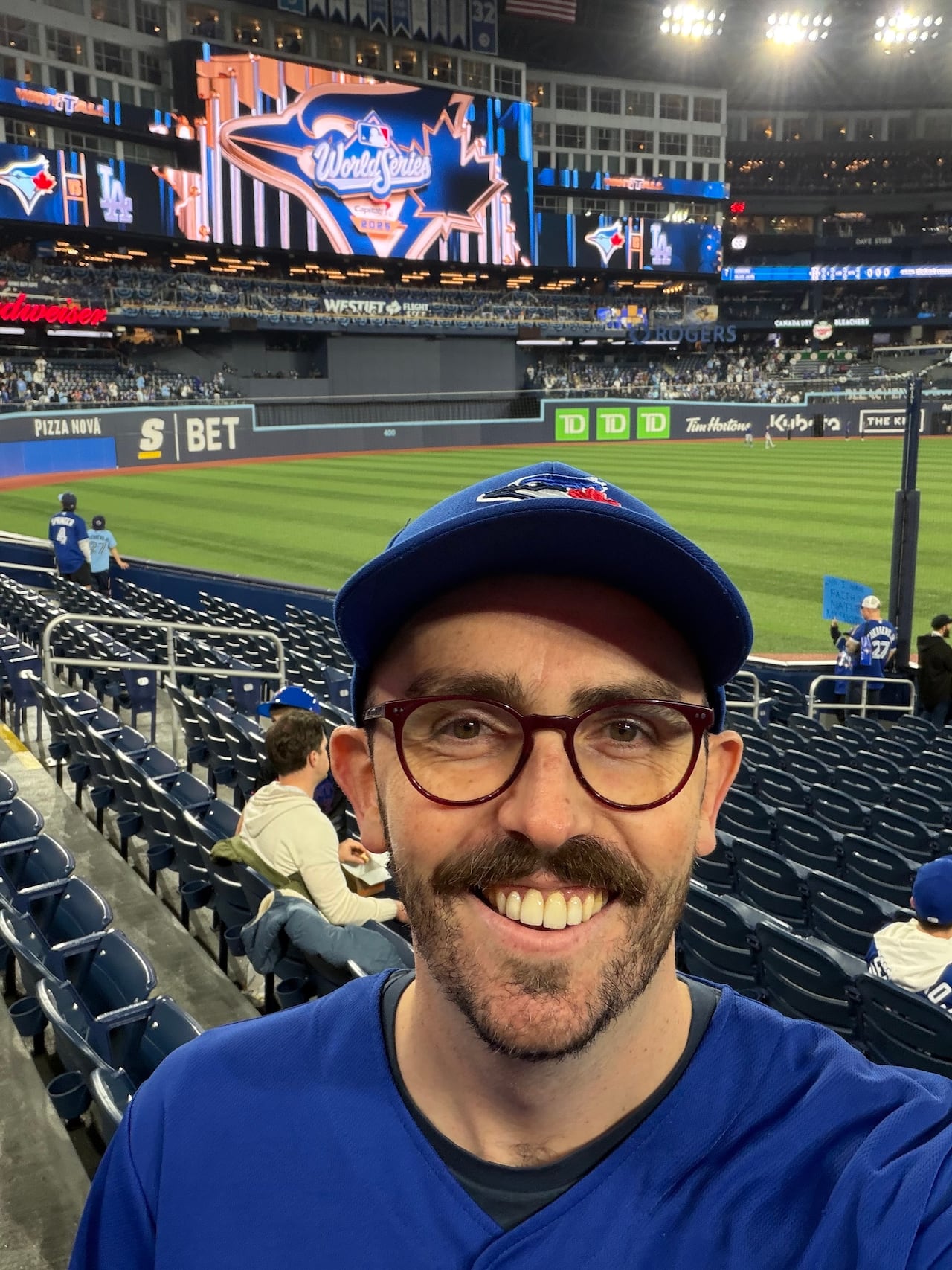 A man with a mustache, wearing glasses and a Blue Jays cap, taking a selfie with a baseball diamond in the background