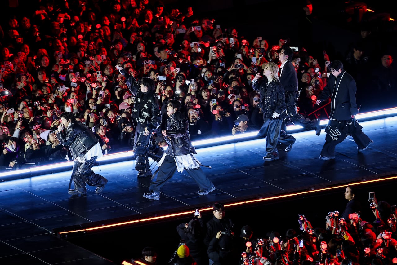A pop group wearing black clothing sings onstage during a concert while attendees take photos. 