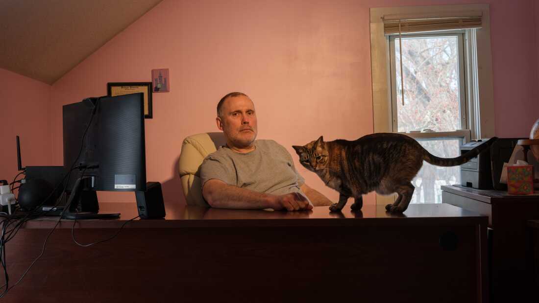 Ken Warner is pictured at his desk, a large computer monitor is visible. His cat is on the desk, looking at the camera. 