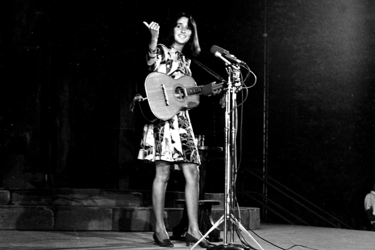 a woman in a knee-length dress stands in front of a microphone holding a guitar on stage, speaking to the crowd and pointing out