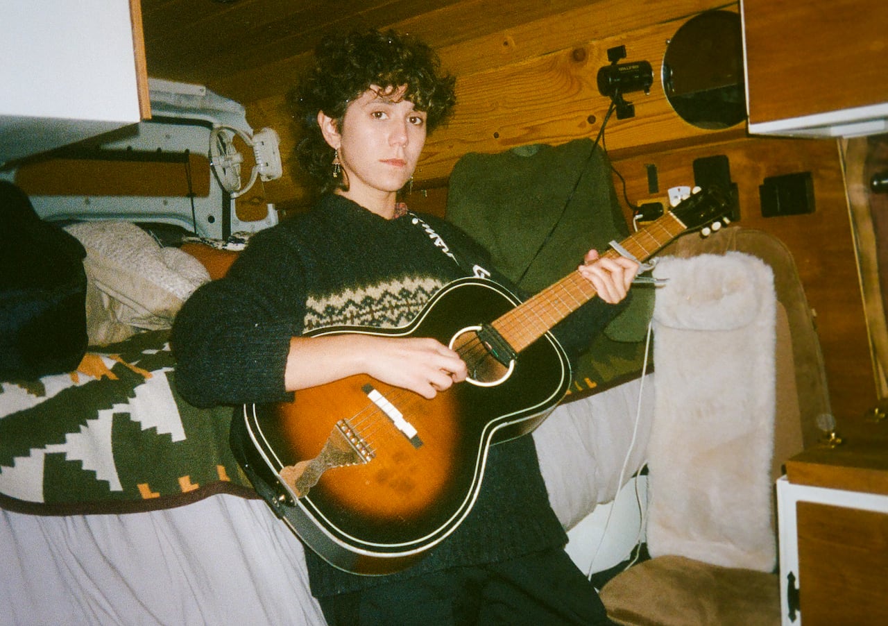 A person with short curly hair wearing a wool sweater leans on a bed and holds an acoustic guitar in their hands