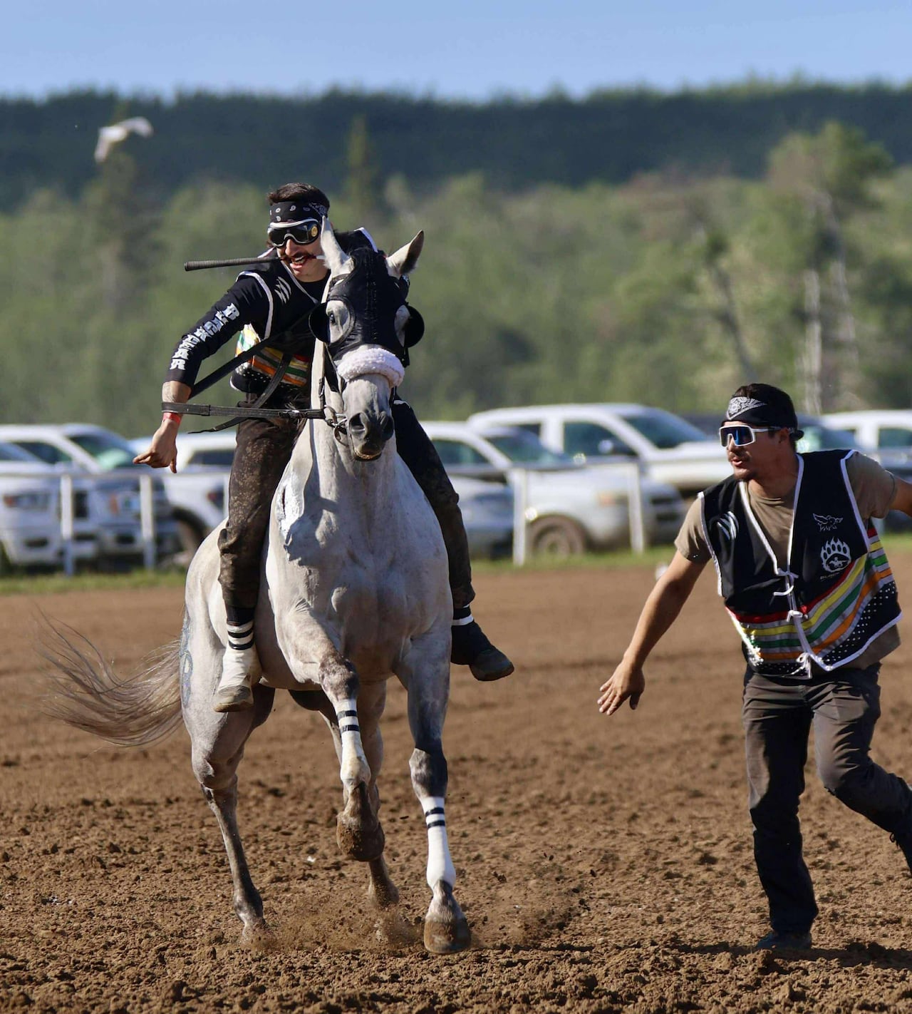 A man rides bareback on a horse with a riding crop in his teeth while another man runs alongside.