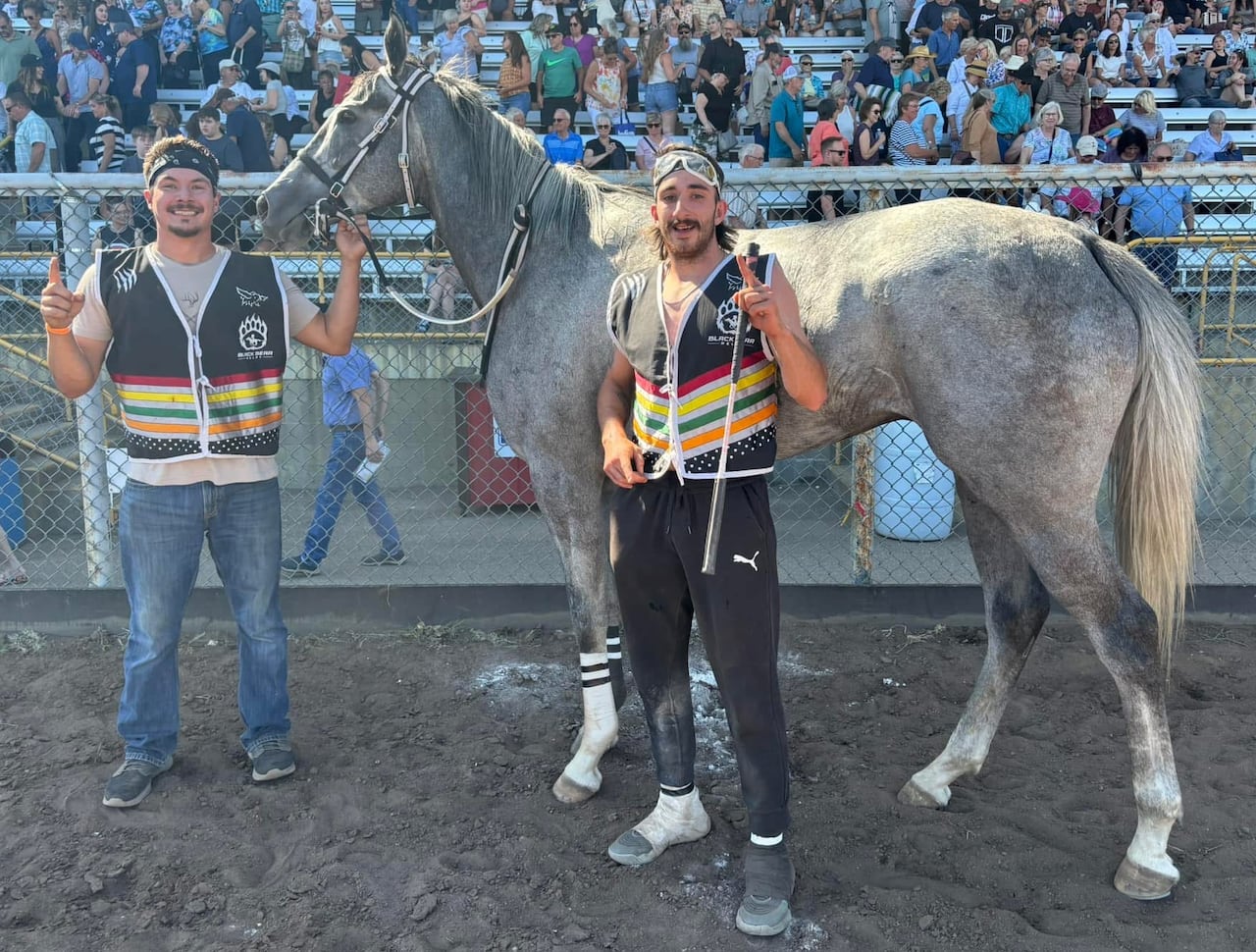 Two men pose for the camera with a grey horse in front of a rodeo audience.