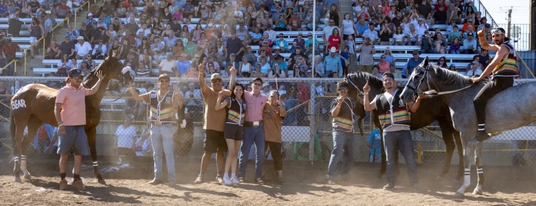 A group of people with three horses gesture at the camera in front of a rodeo crowd. 