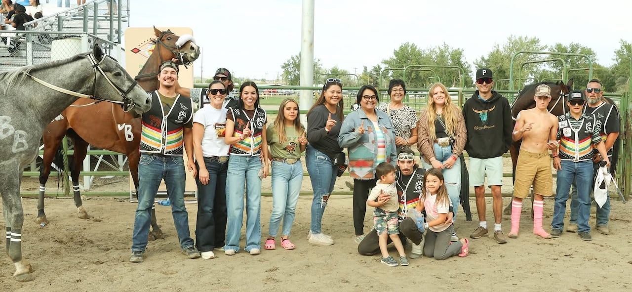 A group of people lined up with three horses smile for the camera on a sunny day at a rodeo ground.