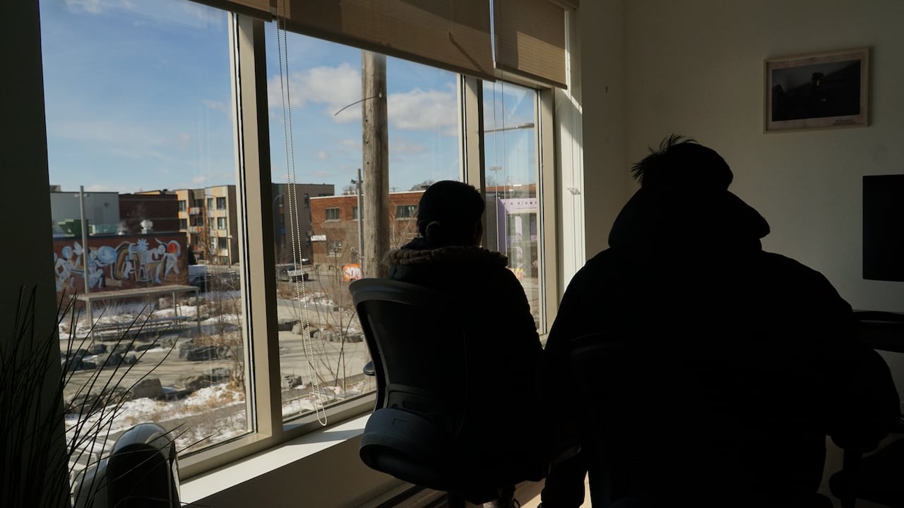 A woman and man sitting at office chairs are seen from behind silhouetted against a window view onto a city park. 