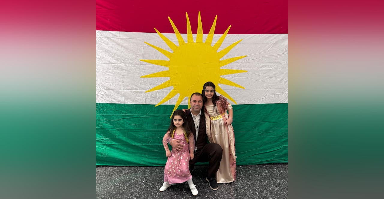 Man with dark hair wears a black suit and hugs two young girls wearing pink dresses infront of a red, white and green flag with a yellow sun
