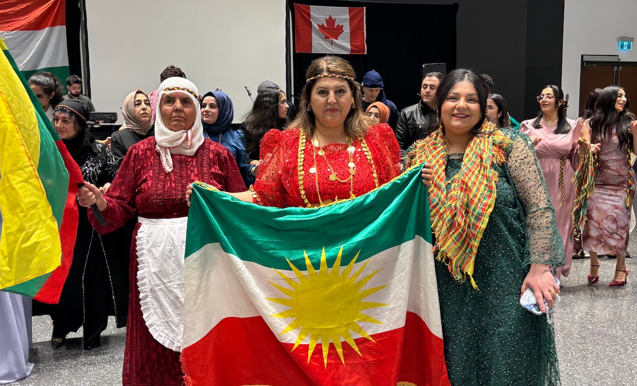 Three women wearing sparkly red and green dresses hold a green, white and red flag with a yellow sun 