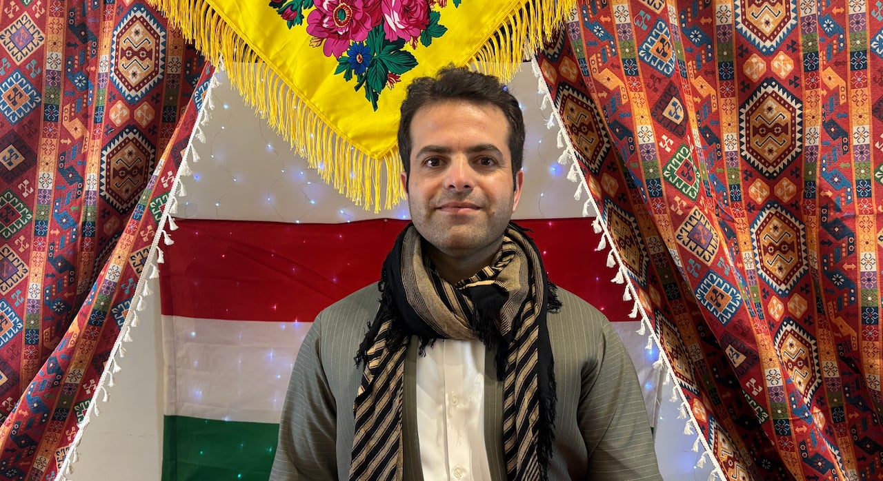 Man with dark hair and dark eyes wears a grey suit with a yellow and black scarf. He stands infront of a colourful display of drapes