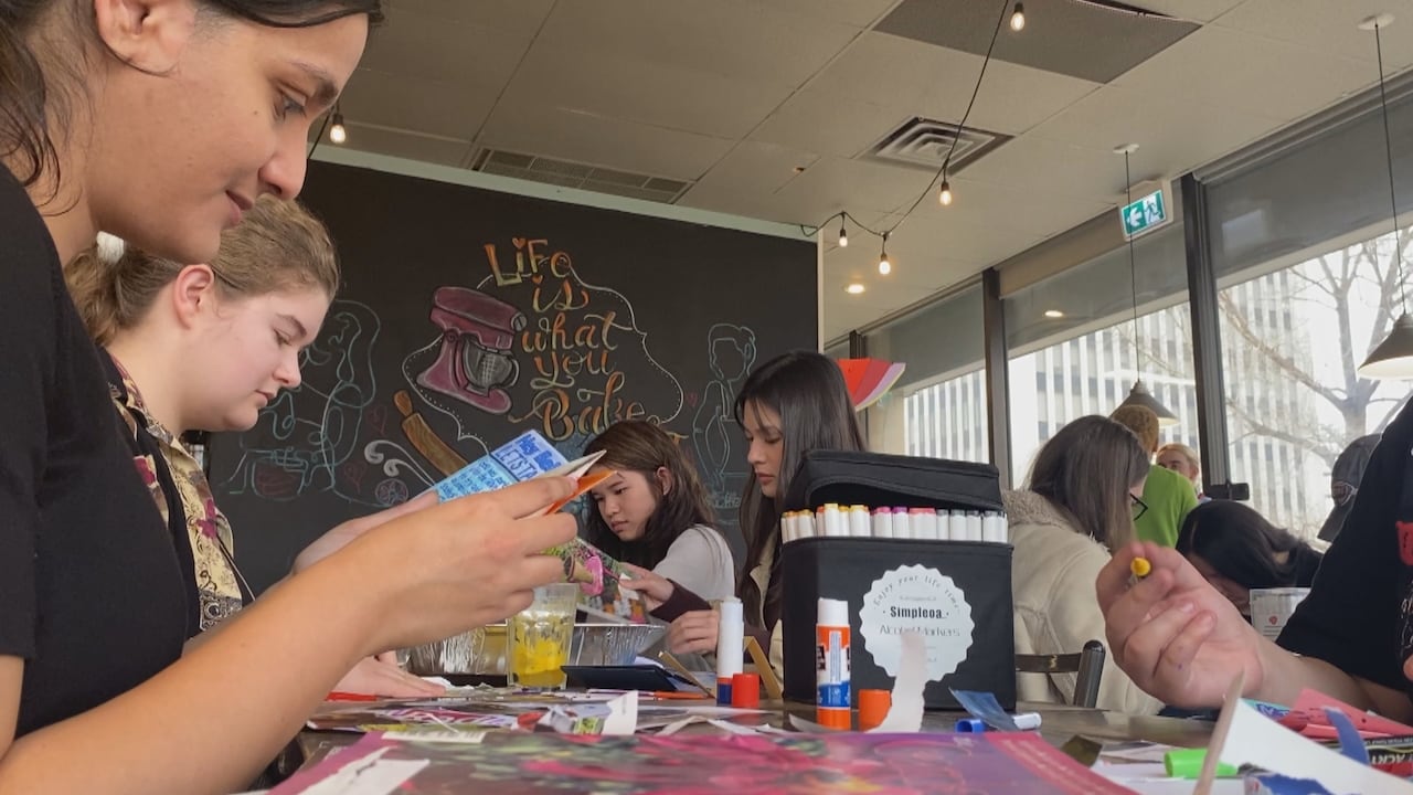 Several young women craft at a table strewn with art supplies as one reads a hand-written pamphlet. 