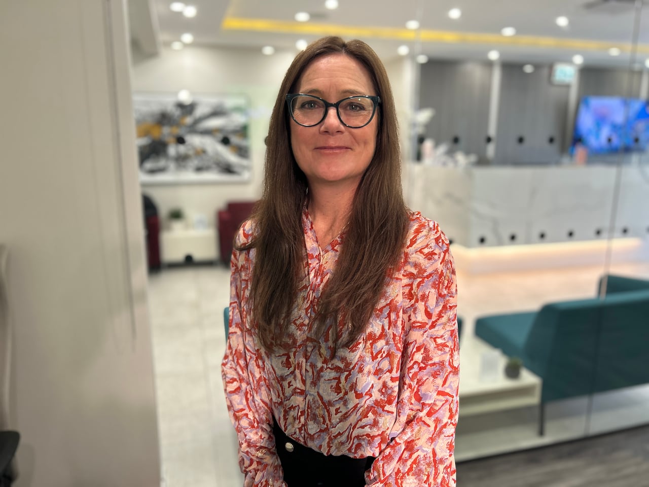 A woman with glasses and long brown hair wearing red blouse stands in a board room.