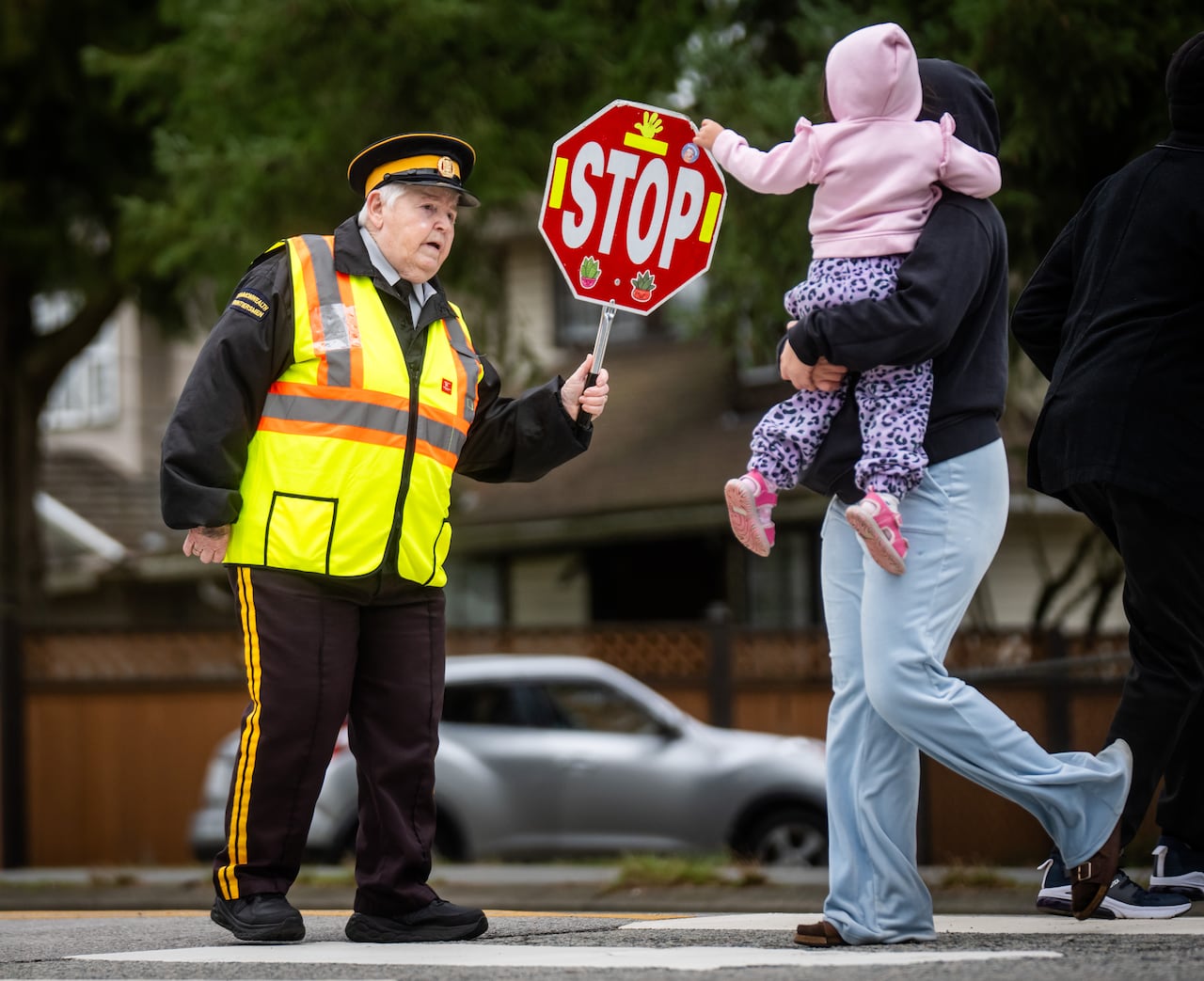 A woman in a neon vest holds a stop sign as an adult holding a child walk by