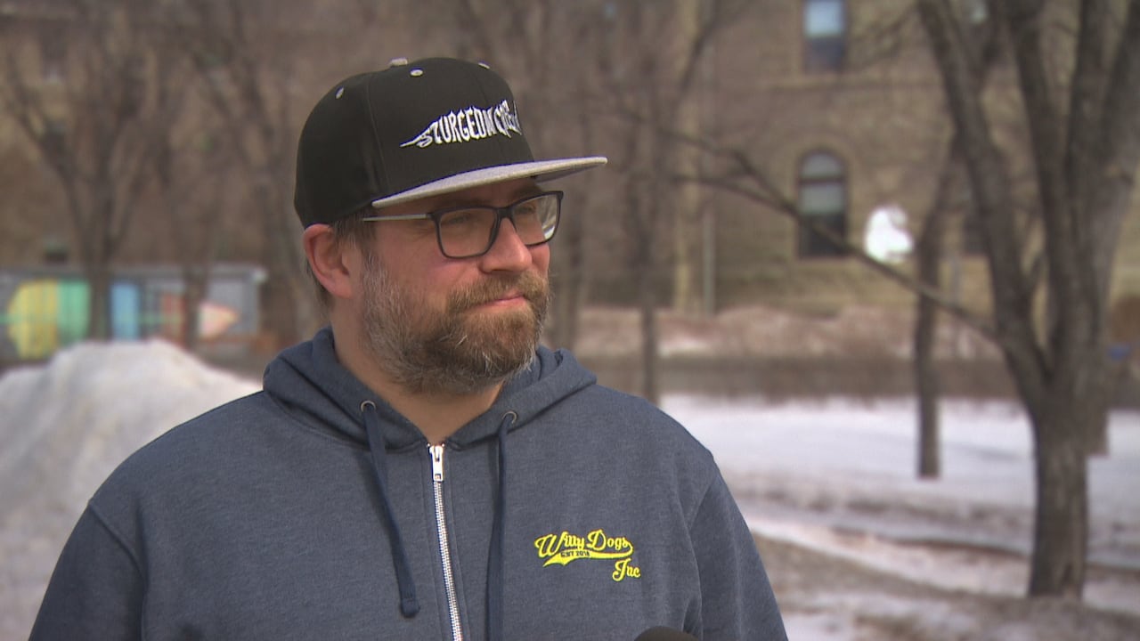 Man with light beard wears eyeglasses and a navy hooded sweatshirt with yellow logo and a black baseball cap