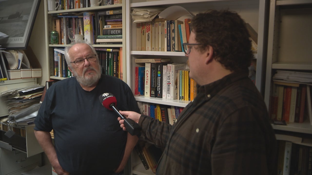 A man in a blue shirt and glasses stands in front of a bookshelf during an interview with a reporter.