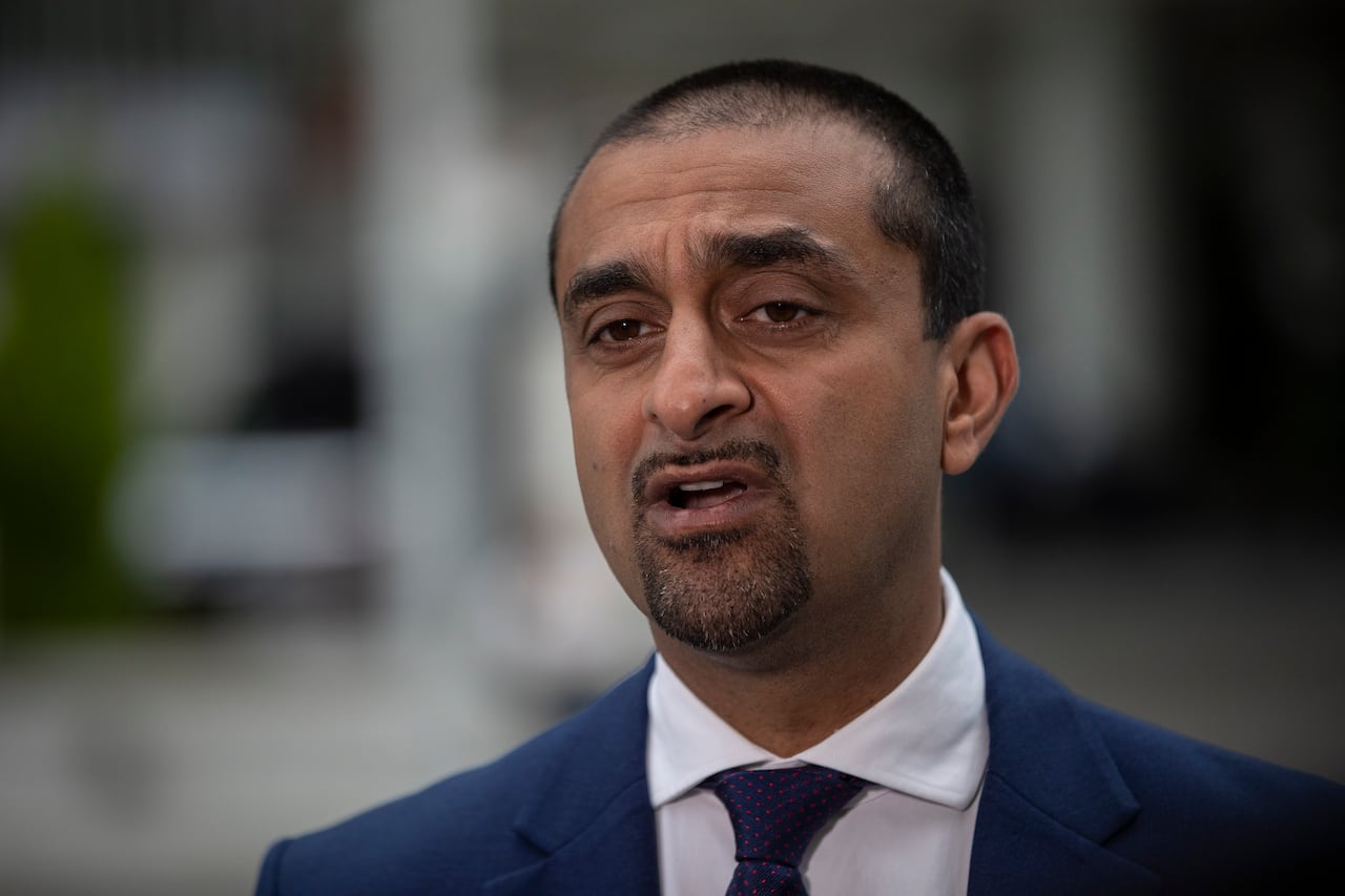 A man with short black hair wearing a suit speaks in a close-up photo.