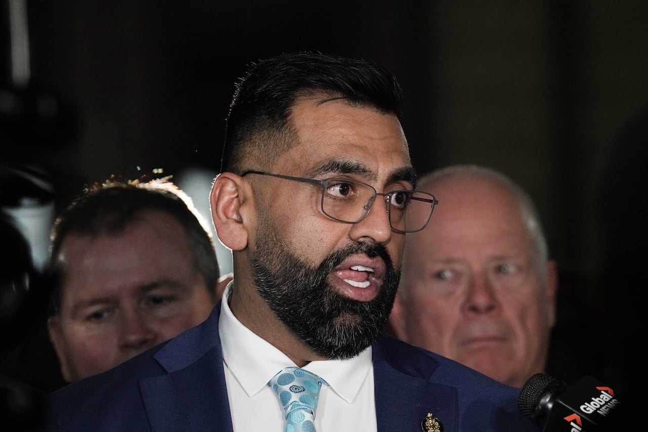 A man in a blue suit, with a dark beard and short dark hair, speaks into microphones during a media interview.