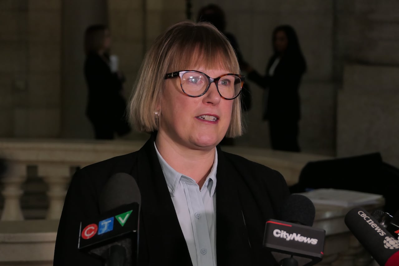 A woman in a black blazer speaks in front of microphones.