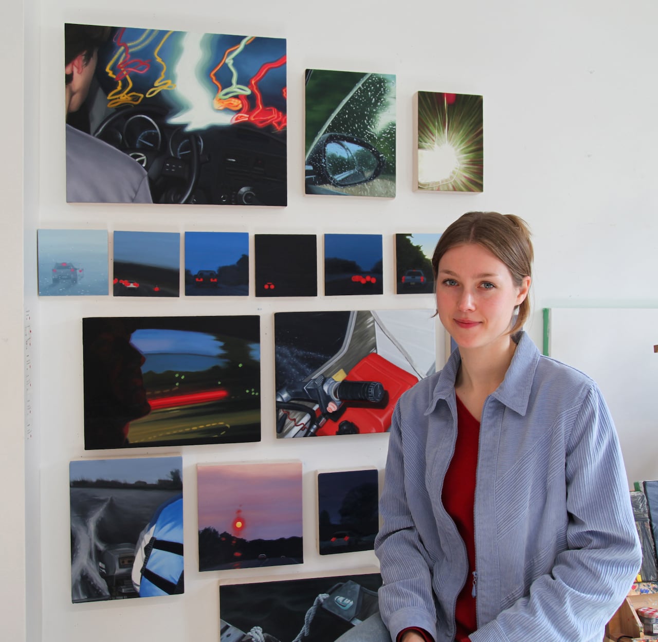Medium shot of the artist Haley Meyer, a woman with blonde hair wearing a blue long-sleeve shirt who is seated in front of a wall of her realistic oil paintings.