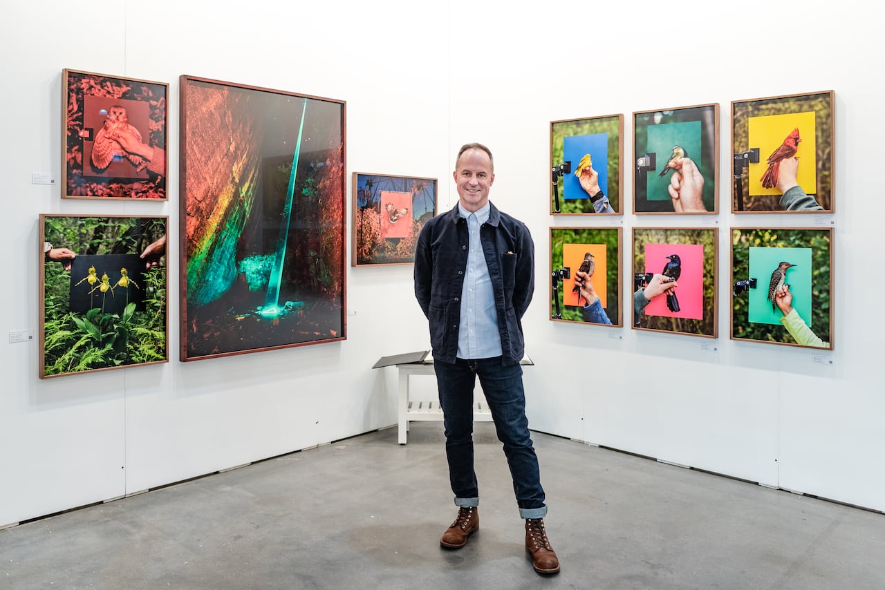 Artist Finn O'Hara is photographed in a full shot at the Artist Project art fair. He is a man wearing a denim-on-denim outfit. He stands, smiling, in front of his photographs which are large in scale and hang on white walls.
