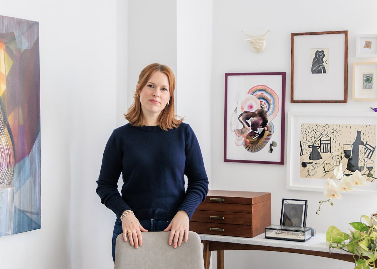 Medium shot of Emily-Jean Alexander, director of Artist Project. She is a woman with red hair wearing all navy blue. She stands behind a chair. Behind her are white walls covered with artwork.