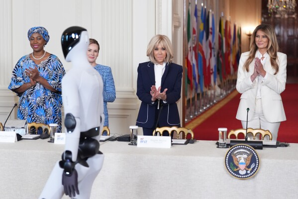First lady Melania Trump arrives, accompanied by a robot, to attend the "Fostering the Future Together Global Coalition Summit," with other first spouses, at the White House, Wednesday, March 25, 2026, in Washington. From left are Dr. Fatima Maada Bio, of Sierra Leone, Martha Nawrocka of Poland, Brigitte Macron of France and Melania Trump. (AP Photo/Jacquelyn Martin)