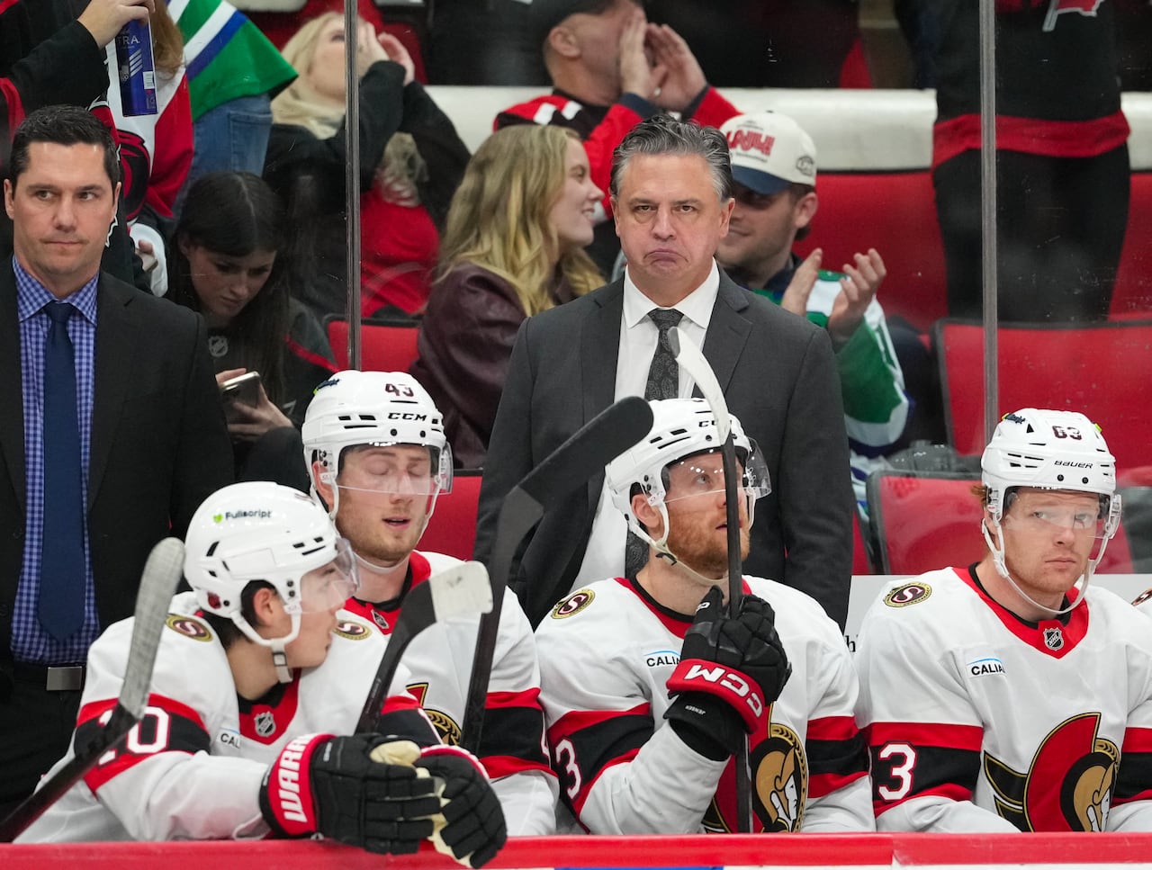 Two coaches and a handful of players watch a hockey game from the bench.