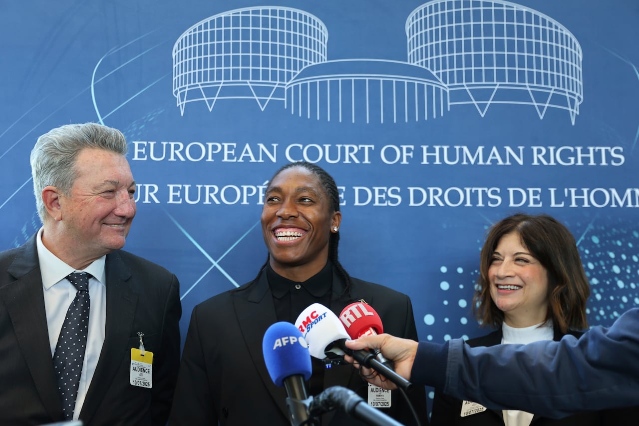 A person smiles into a microphone flanked by two people in suits