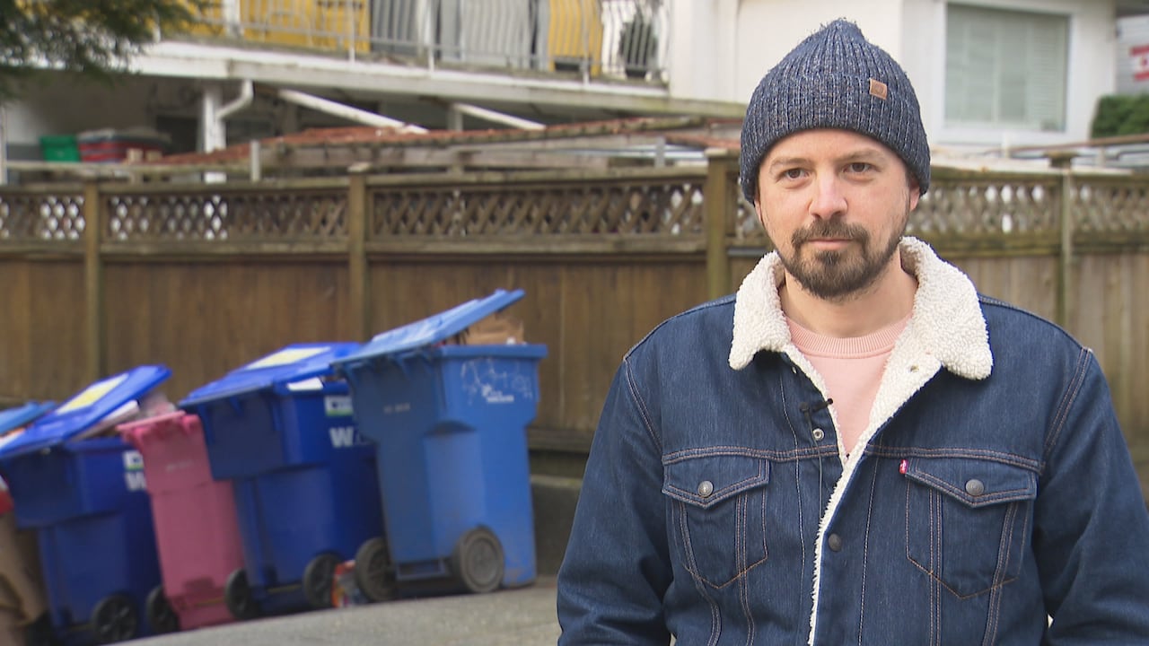 A man in a jean jacket and tuque is seen outside, with a row of full recycling bins behind him.