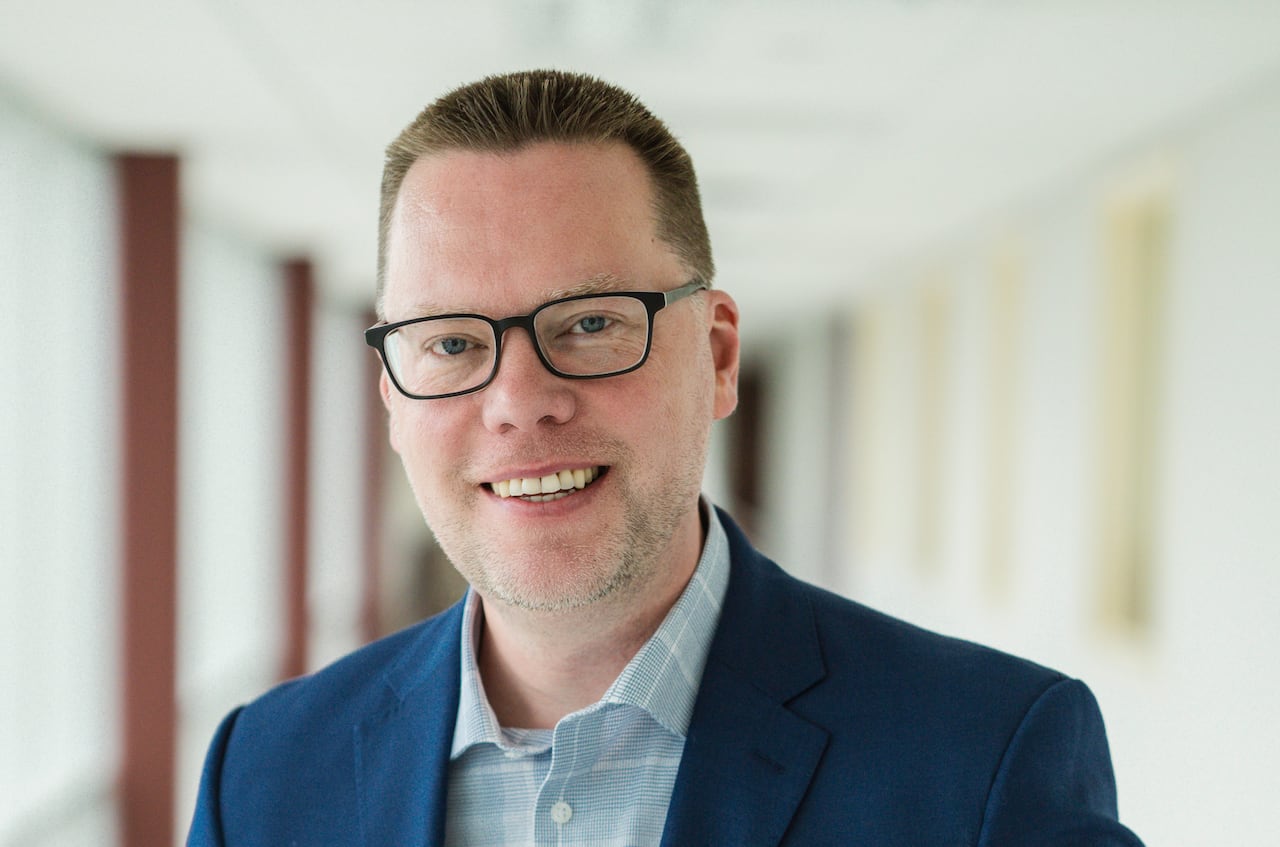 A professional portrait of a man with short brown hair and glasses, wearing a blue jacket and checked shirt.