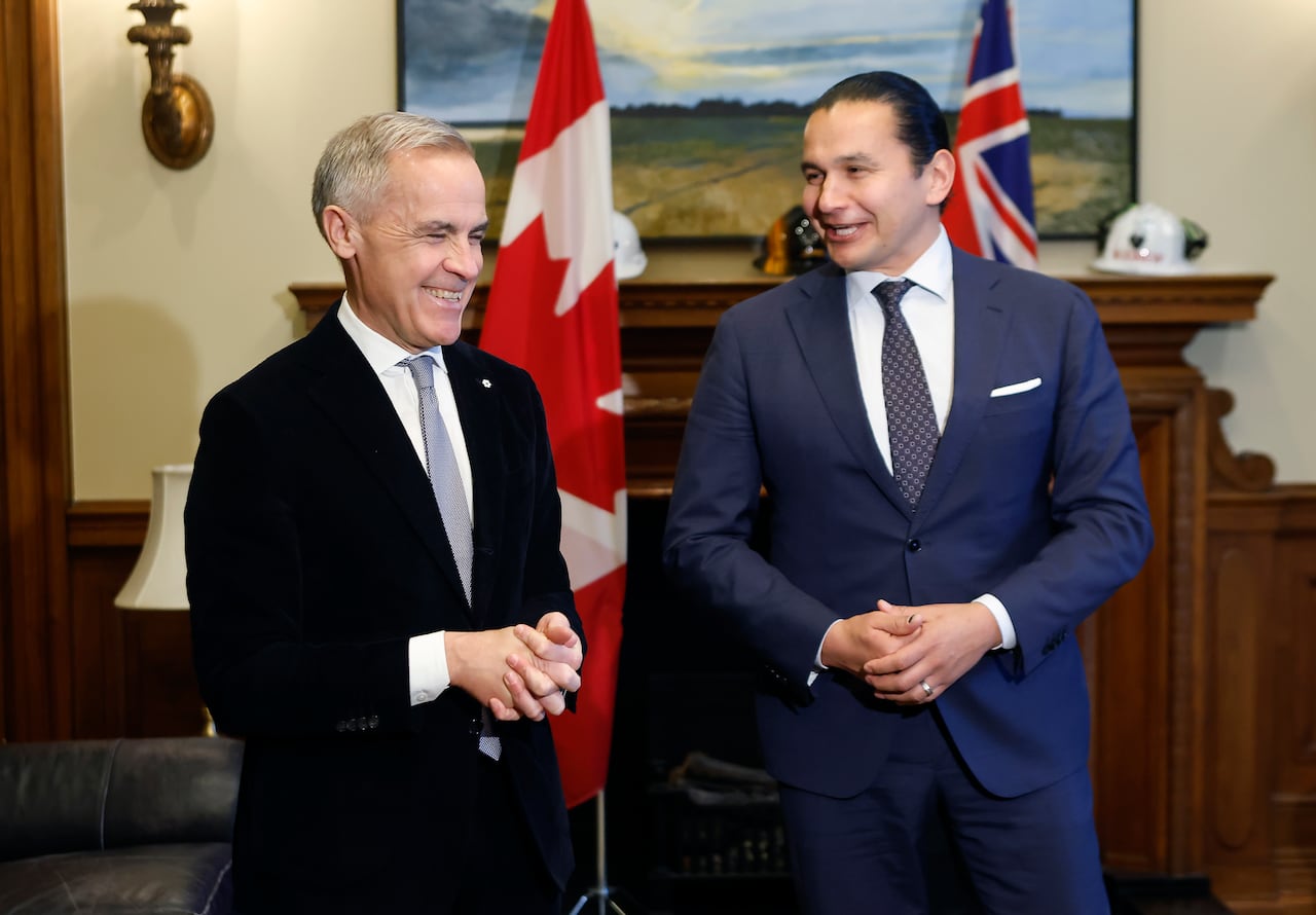 Two men in suits smile as they stand next to each other in a room with Canadian and Manitoba flags.