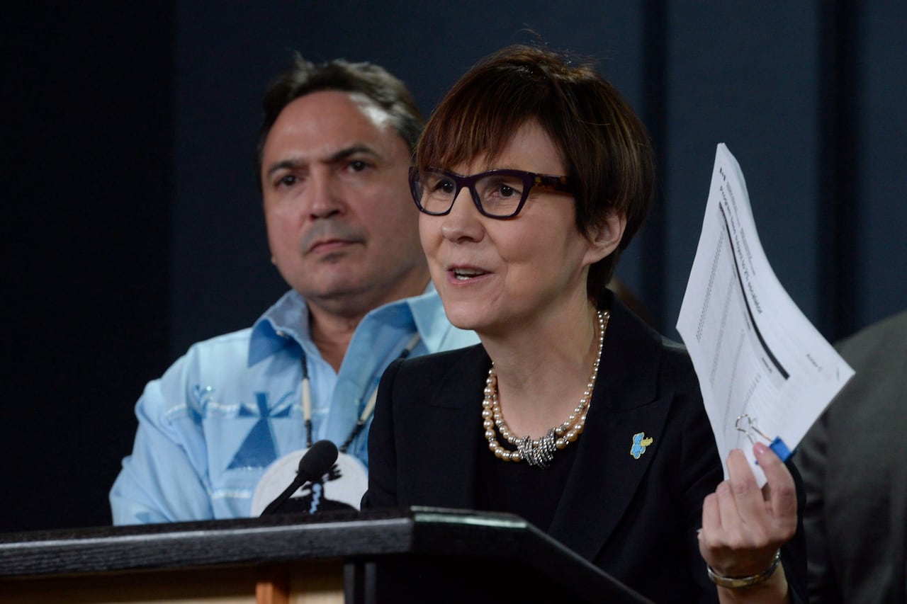 A woman in black glasses and black clothing speaks at a podium. A man in a blue shirt sits behind her.
