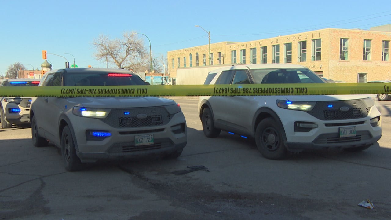 Several police cars are shown with flashing red and blue lights behind a line of yellow police tape along a city road. 