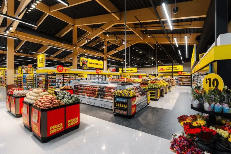 An interior view of a modern grocery store showcasing fresh fruits, vegetables, and flowers, with bright lighting and clear signage for discounts and promotions.