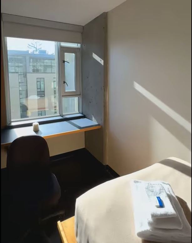 A small dorm room with a bed, folded towels, a desk and chair by a window, and sunlight coming through.