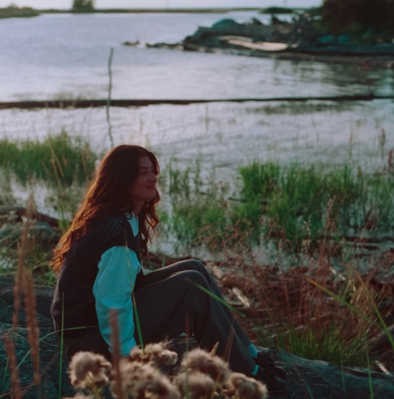 A woman sits among grass and rocks near a body of water